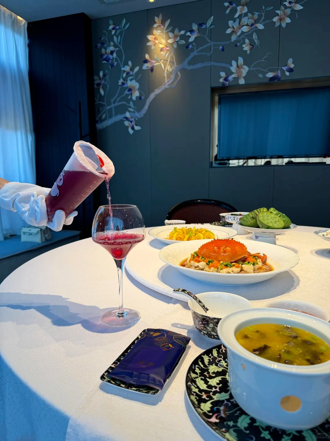 A gloved hand pours a Mixue cup into a wine glass on a banquet table set with crab, soup and lotus, under a painted wall panel