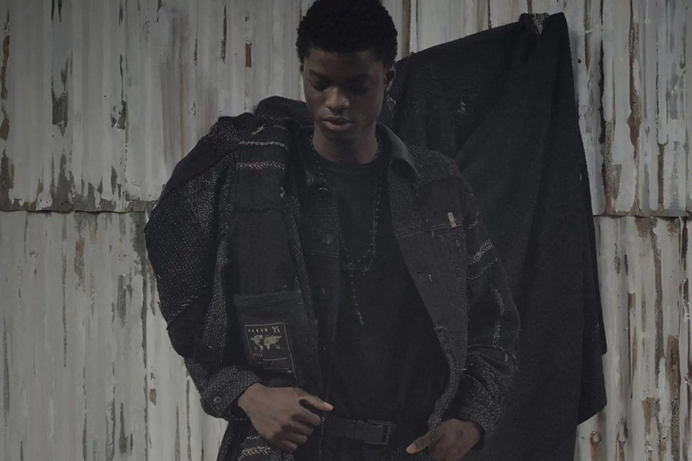 A black coat with visible kantha quilt stitching hangs against a neutral background, part of the Y's and Fake As Flowers capsule collection